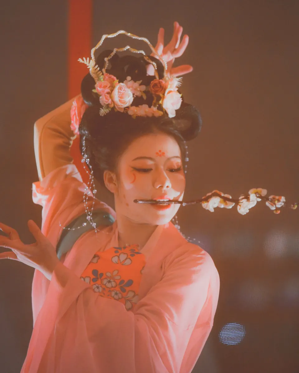 A dancer in a pink Tang-style court costume and floral headdress, holding a blossom sprig between her lips on stage