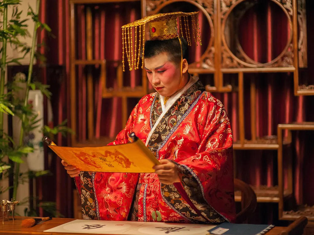 A performer in an emperor-style court robe and beaded crown reading a scroll in a room of carved wooden shelves